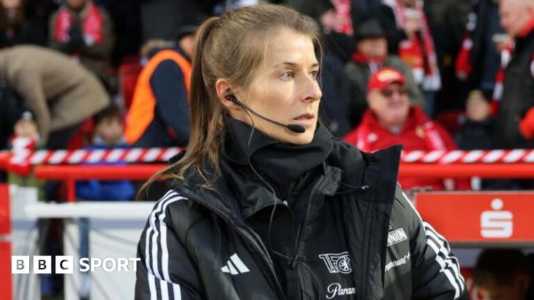 Union Berlin appoint Marie-Louise Eta as first woman head coach Marie-Louise Eta, wearing a black coat and top with a black microphone and headset in her right ear, on the touchline with fans in red and dark clothing in the background