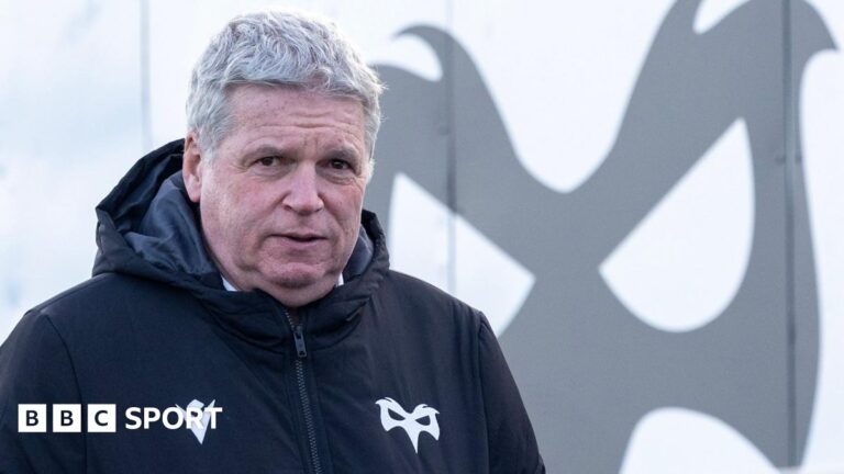 Lance Bradley wearing an Ospreys jacket in front of an Ospreys logo at Brewery Field