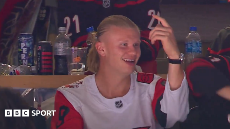 Erling Haaland, wearing a white Carolina Hurricanes jersey with red, black and white sleeves, smiles and points his left hand in the air