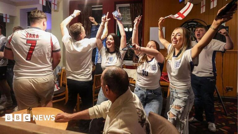 A group of men and women wearing England shirts - with one waving an England flag - celebrate in a pub, with their arms in the air