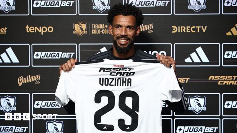 Cape Verde’s Vozinha granted shirt name exemption by Chile FA Vozinha, smiling and holding a white Colo Colo shirt with Vozihna 29 on the back in front of a black board of sponsor logos